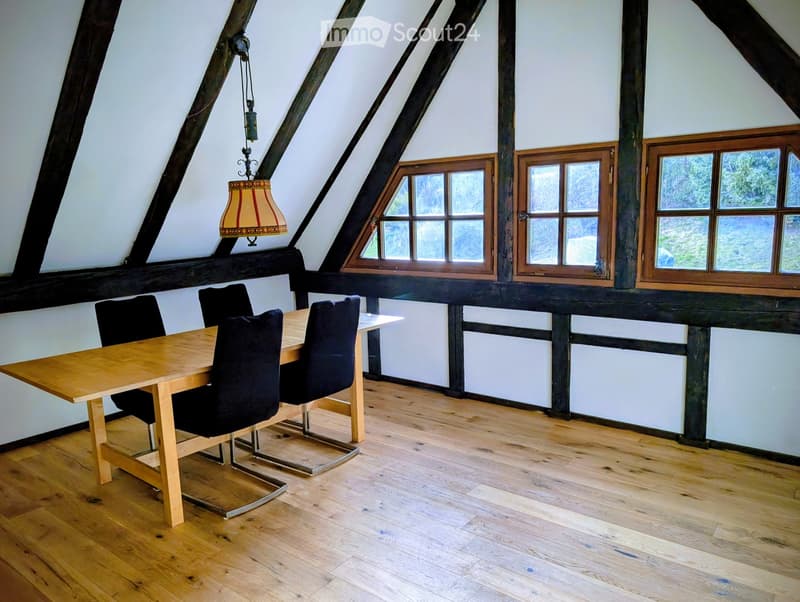 Bright attic room with wooden floors, exposed wooden beams, large windows, a dining table with chairs, and a hanging lamp.