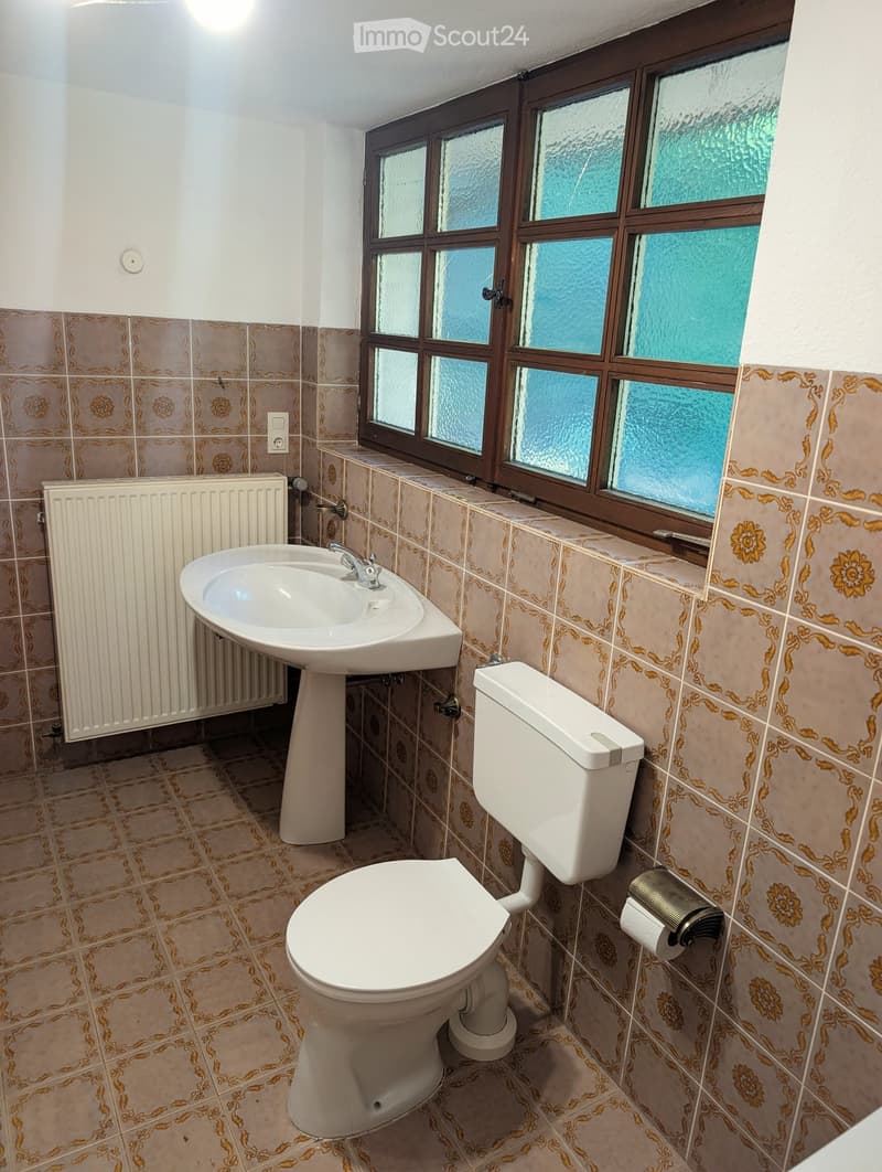 Bathroom with beige tiles, a toilet, a sink with a faucet, a radiator, and a toilet paper holder. The window has frosted glass panes.