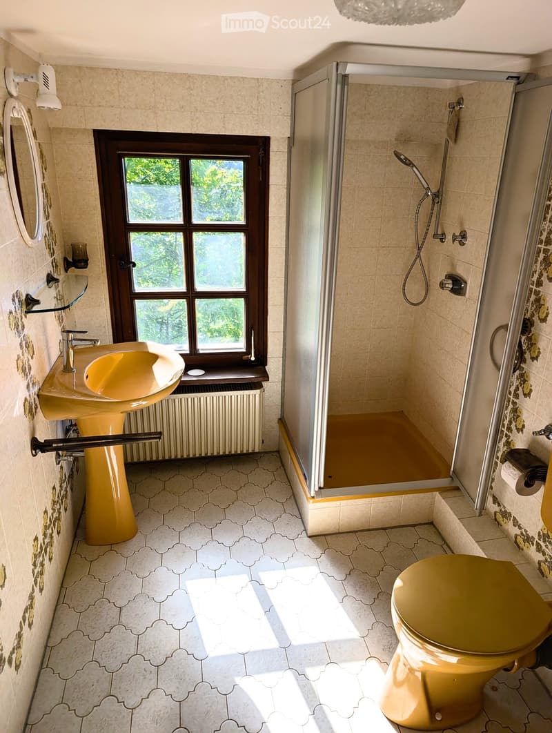 Bathroom with a yellow sink, matching toilet, glass shelf, and a shower with a handheld sprayer. The room features patterned tiles and a window with a view of greenery.