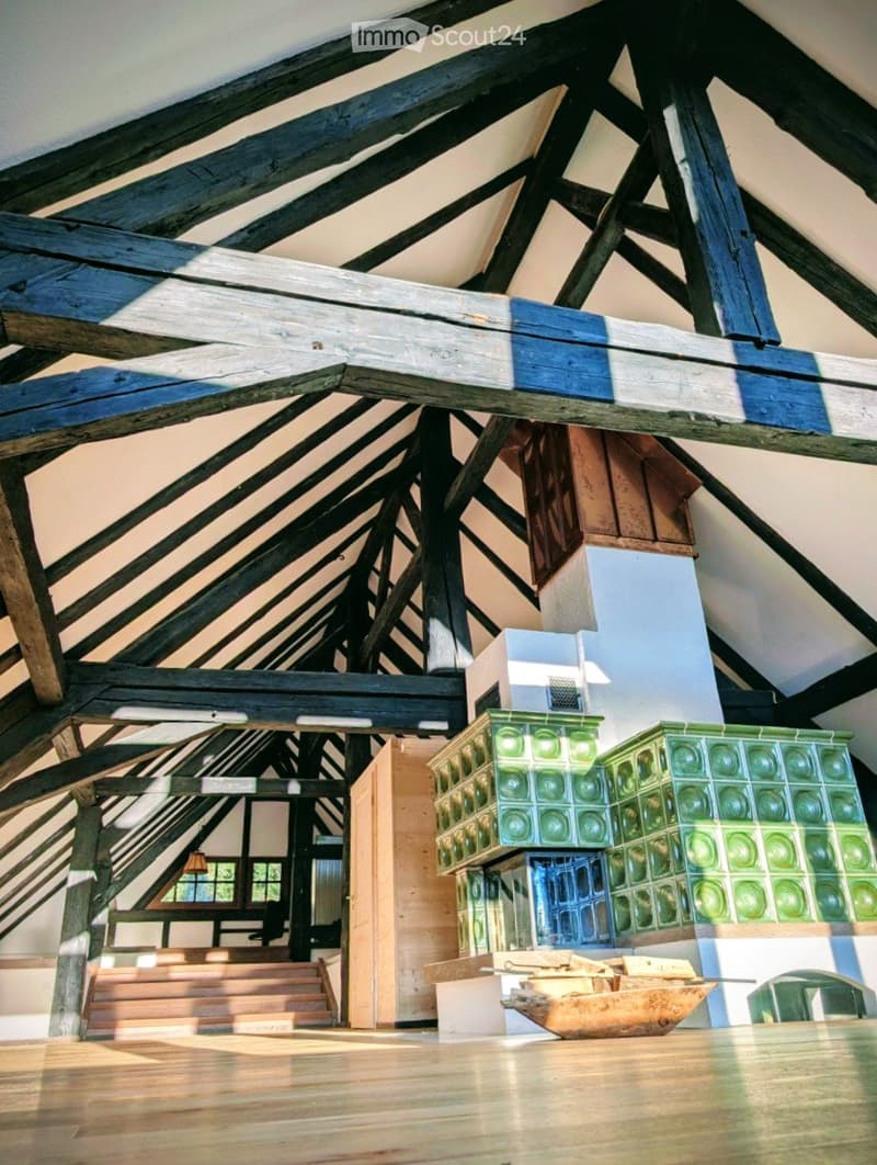 An attic space with exposed wooden beams and a sloped ceiling, featuring a wooden floor, a staircase leading up, and a small landing area with glass block walls.