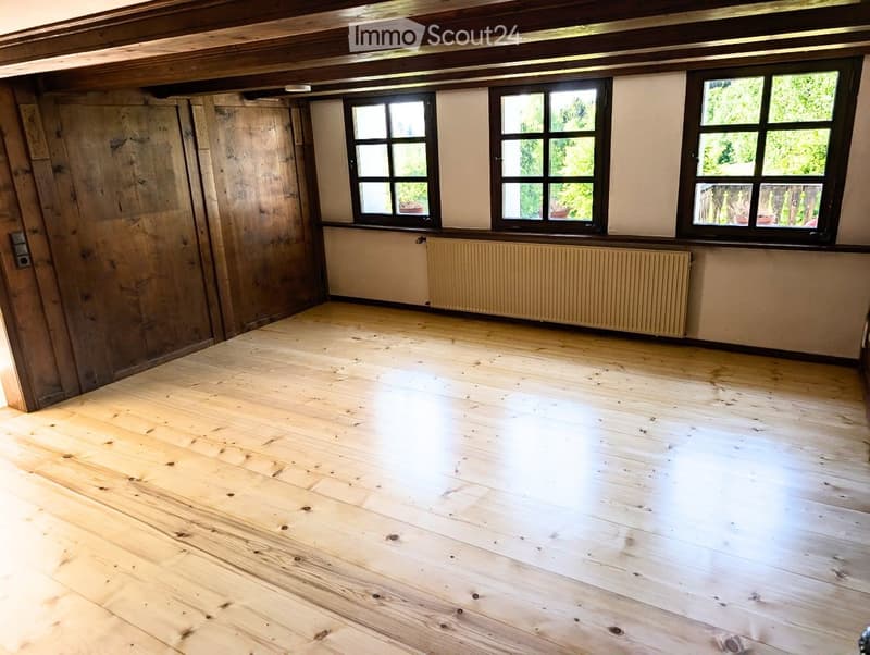 Empty room with wooden floor, exposed wooden beams on the ceiling, and three windows with black frames. The walls are a mix of wood paneling and white plaster.