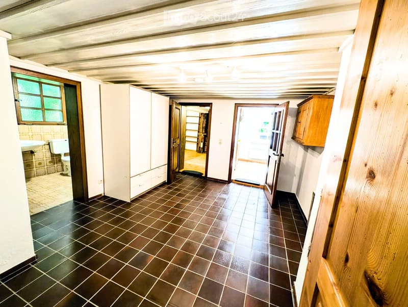 A narrow hallway with dark tiled flooring, white walls, and wooden beams on the ceiling. There are several doors leading to different rooms, including a bathroom and a closet. The hallway is well-lit with natural light coming from a window on the left sid