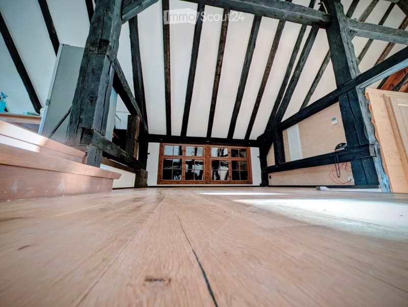 Empty attic space with exposed wooden beams, wooden floor, and a small wooden cabinet with glass doors.