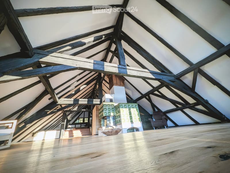 Loft-style room with exposed wooden beams and a high, slanted ceiling. The space features a wooden floor, a brown sofa, and a small table with a patterned top.