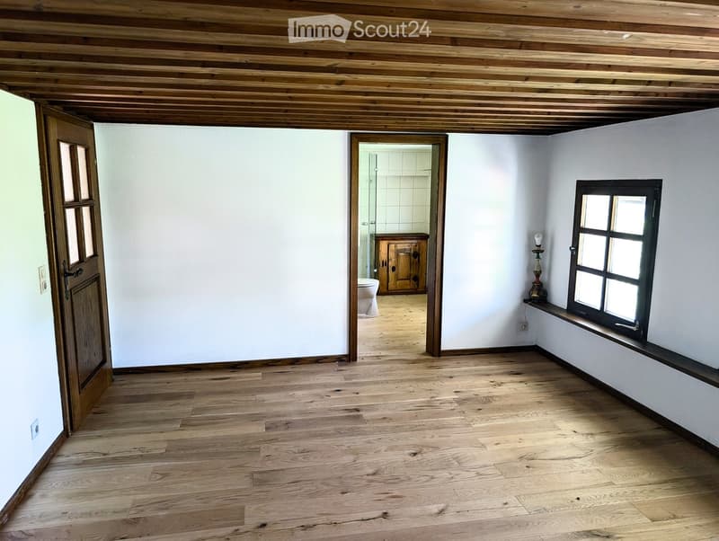Empty room with wooden flooring, white walls, and a wooden ceiling. Features a window with a small shelf and a doorway leading to a bathroom.