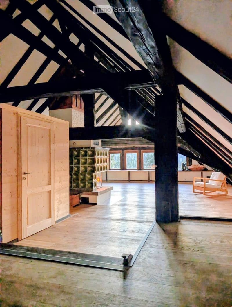 Spacious attic room with exposed wooden beams, large windows, polished wooden floors, and modern furniture including a chair, table, and stool.