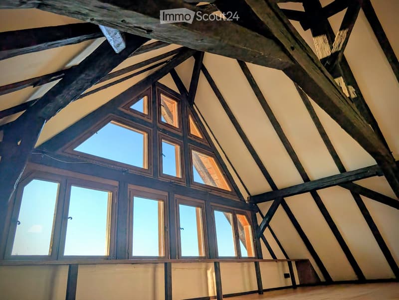 An attic space with exposed wooden beams and dormer windows, featuring a wooden floor and light-colored walls.