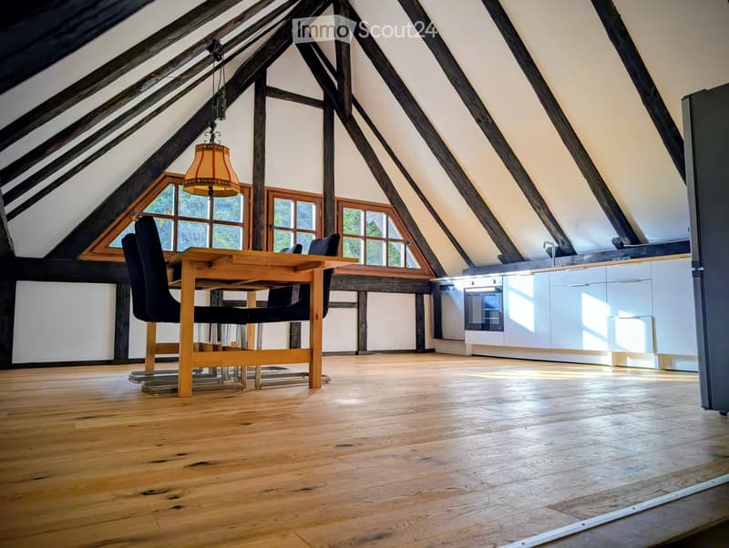 Open-plan attic space with wooden floors, exposed wooden beams, large windows, a dining area with a table and chairs, and a modern kitchen with white cabinets and stainless steel appliances.