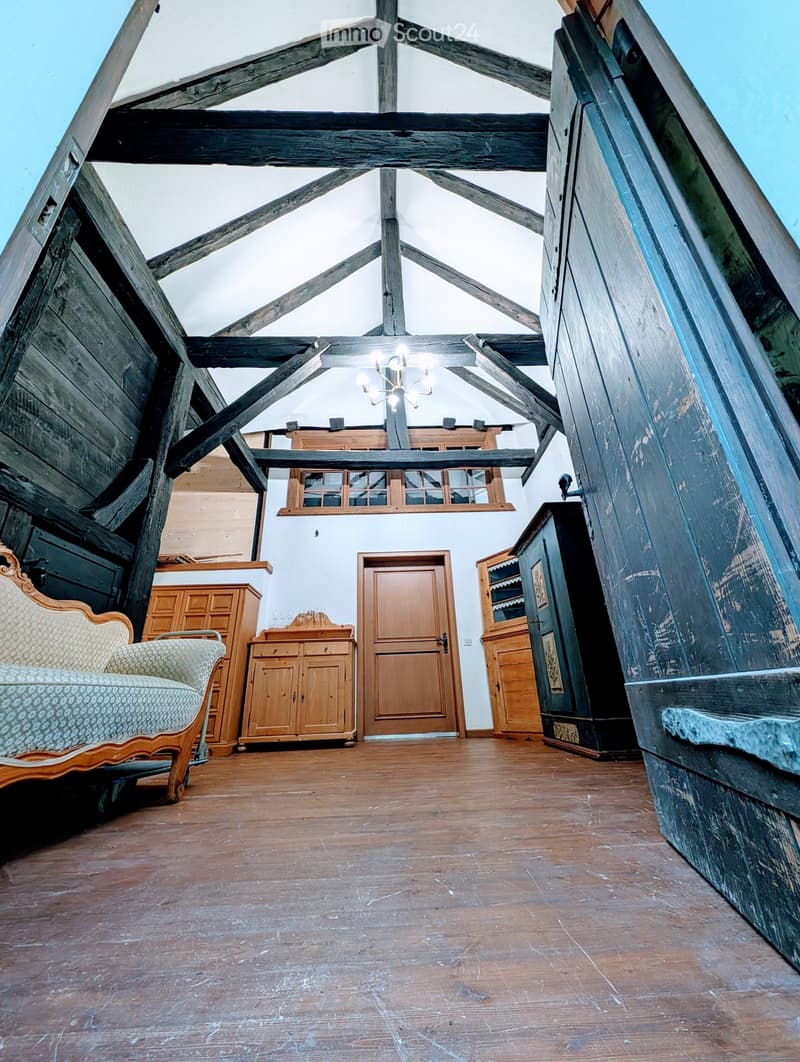 A spacious, high-ceilinged room with exposed wooden beams, wooden flooring, and a mix of antique and modern furniture including a patterned sofa, wooden cabinets, and a chandelier.
