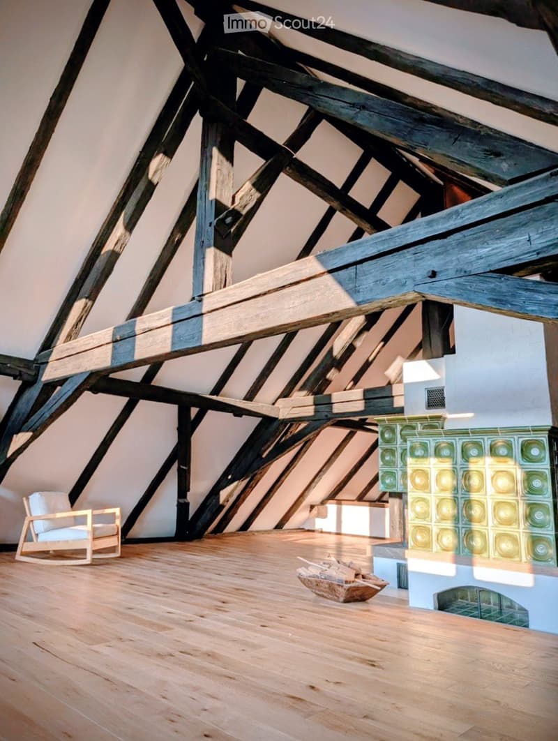 An attic space with exposed wooden beams, a wooden floor, a white chair, and a fireplace with a decorative tile surround.