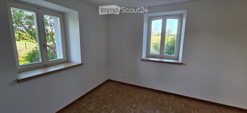 Empty room with wooden flooring, white walls, and two windows with wooden sills, offering a view of greenery outside.