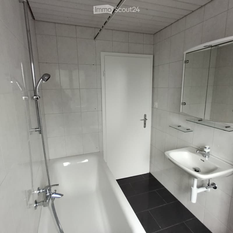 Modern bathroom with a bathtub, shower, sink, and mirror. The walls are tiled in gray, and the floor is black.