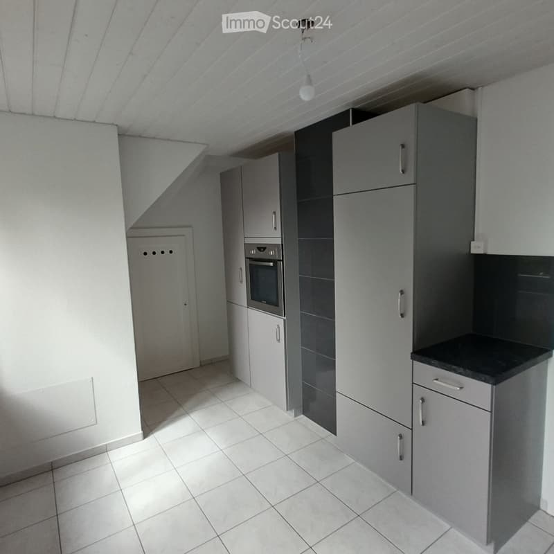 Modern kitchen with grey cabinets, white tiled floor, and built-in oven. The space is clean and well-lit with natural light.