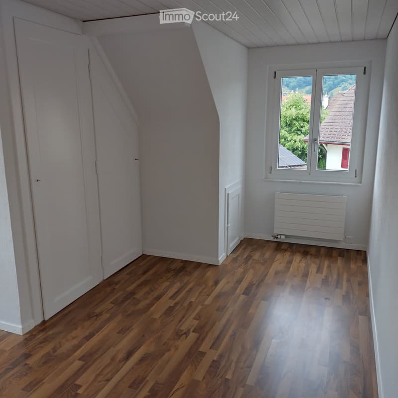 Empty attic room with wooden flooring, white walls, a window with a view, and a radiator. No furniture or equipment present.