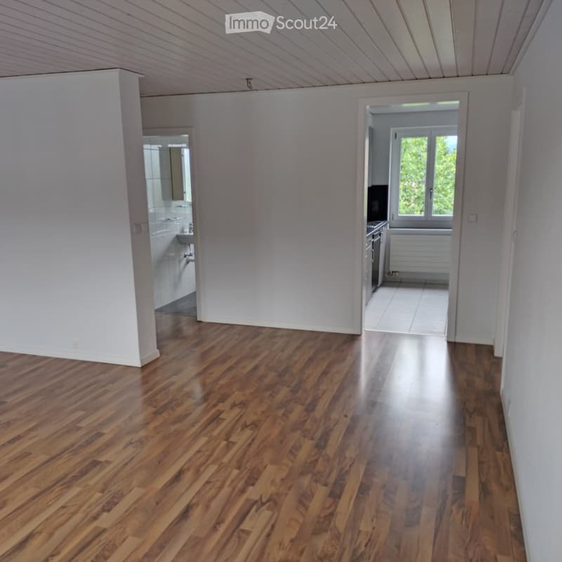 Empty hallway with wooden flooring, white walls, and open doorways leading to a bathroom and kitchen area.