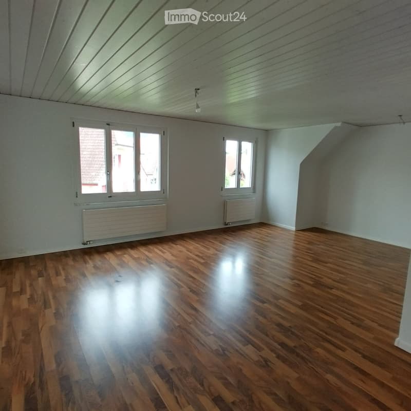 Empty room with wooden flooring, white walls, and large windows, featuring radiators and a slanted ceiling.
