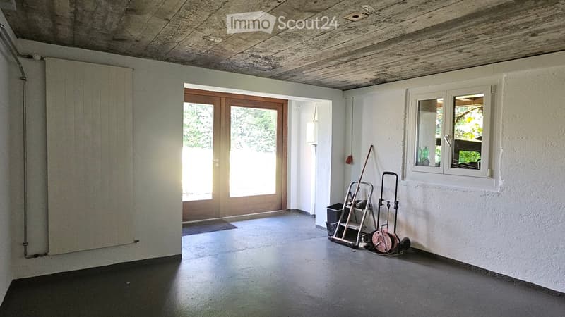 Empty room with white walls, concrete ceiling, large wooden door, and a window with a view of greenery. A small cart with tools is placed near the window.