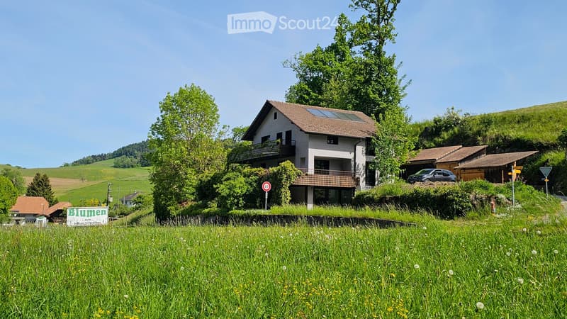 Two-story house with a balcony, surrounded by green fields and trees. The house has a brown tiled roof and is partially covered in greenery. A car is parked nearby, and there is a speed limit sign visible.