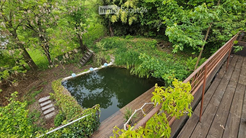 A serene garden area with a small pond surrounded by lush greenery, wooden decking, and a metal railing. Stairs lead down to the pond, and the area is enclosed by trees and plants.
