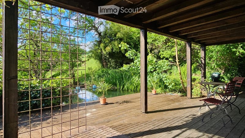 Wooden deck with a table and chair, surrounded by lush greenery and a small pond.