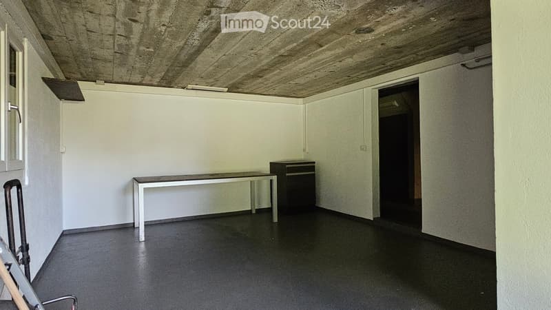 A small, unfurnished room with a concrete ceiling, white walls, and a black floor. A simple table and a cabinet are present, with a doorway leading to another space.