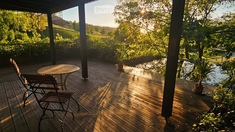 Wooden deck with a round table and two chairs, surrounded by lush greenery and a small water feature.