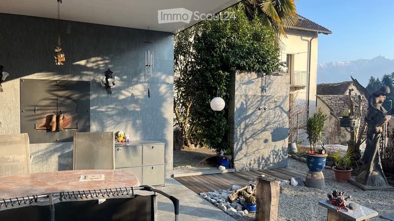 Outdoor patio with a wooden table, chairs, barbecue, potted plants, and decorative items, overlooking a scenic mountain view.