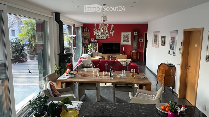 Bright living and dining area with red accent wall, large windows, and a view of the garden. Features a sofa, dining table with chairs, television, chandelier, fireplace, and decorative items.