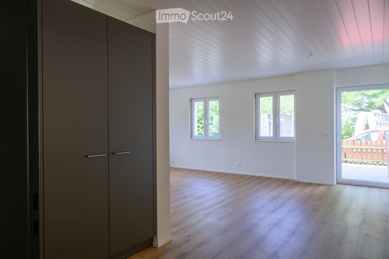 Empty room with light wooden flooring, white walls, and large windows, featuring a sliding glass door leading outside.