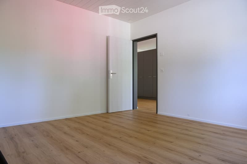 Empty room with light-colored walls and wooden flooring, featuring a closed door and an open doorway leading to another room with dark cabinets.