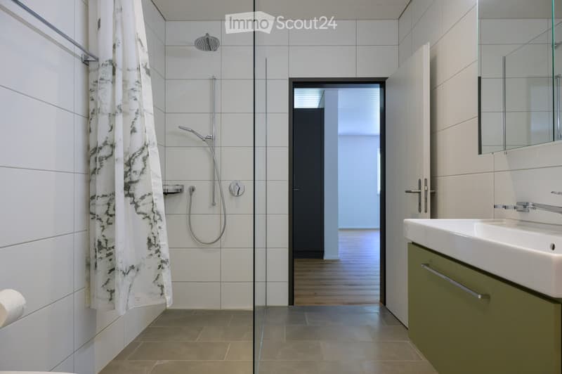 Modern bathroom with white tiles, glass shower enclosure, a sink with a mirror, and a towel hanging on the wall.