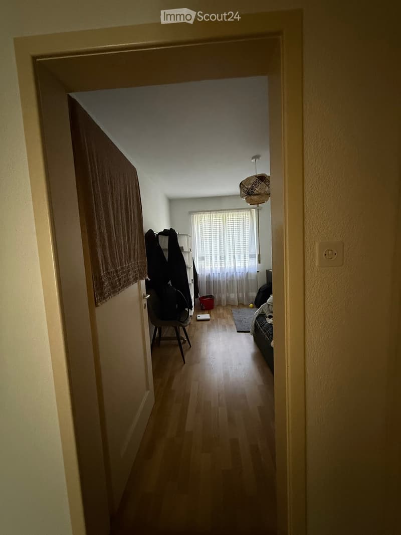 A narrow hallway leads to a furnished room with a bed, chair, and a hanging lamp. The room has wooden flooring, a window with curtains, and a small rug near the bed.