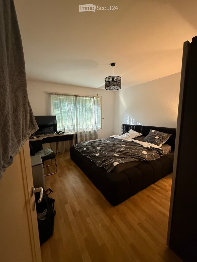 Modern bedroom with wooden flooring, a large bed with dark bedding, a desk with a laptop, and a hanging lamp.
