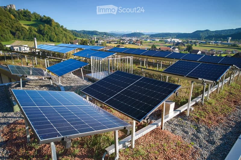 A rooftop solar panel installation with multiple panels mounted on a metal frame, surrounded by a gravel surface and greenery, with a scenic valley and mountains in the background.