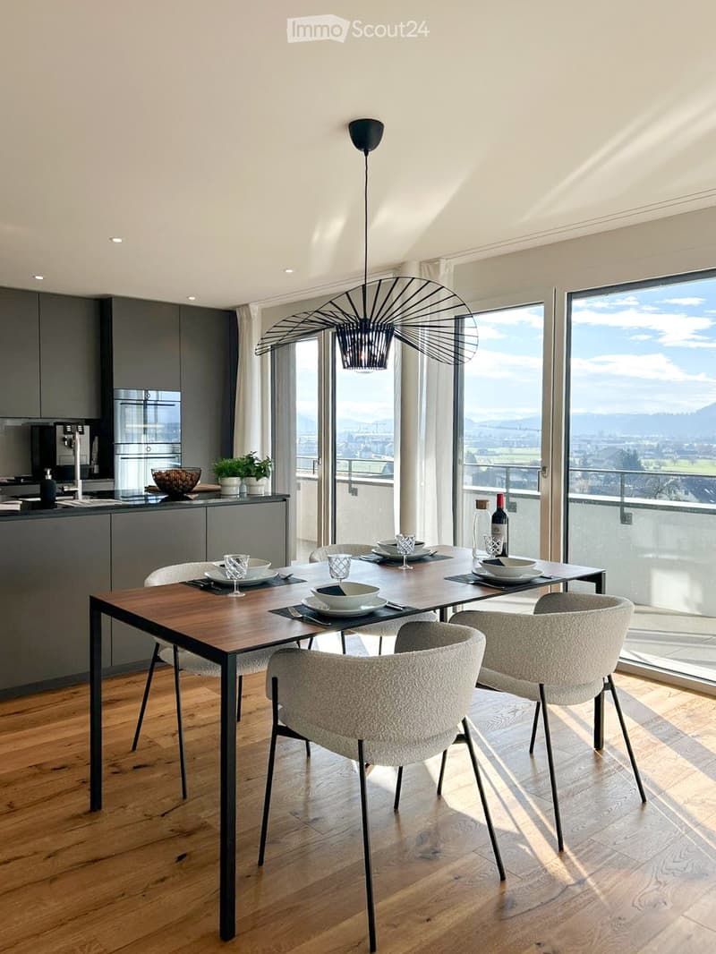Modern dining area with a wooden table, six chairs, and a view of the cityscape through large windows. The area includes a kitchen counter with a refrigerator, potted plants, and a wine bottle on the table.