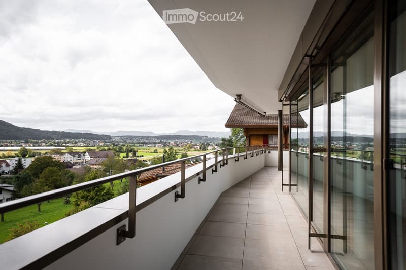 Modern balcony with tiled floor, glass railings, and metal balustrade, overlooking a scenic countryside with houses, trees, and distant mountains.