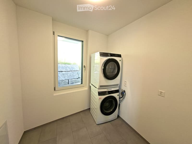 A small, well-lit room with tiled flooring and white walls, featuring a stacked washing machine and dryer unit near a window with a view of a stone wall and greenery.