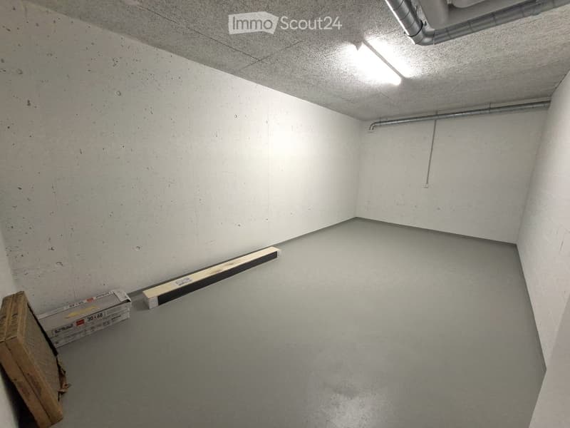 Empty room with concrete walls and ceiling, exposed pipes, and a small window letting in light. There are some storage boxes and wooden planks on the floor.