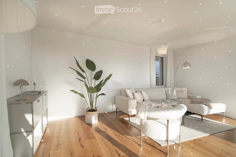 Modern living room with white walls, wooden flooring, a large sectional sofa, a glass coffee table, a white armchair, a potted plant, and decorative lamps.