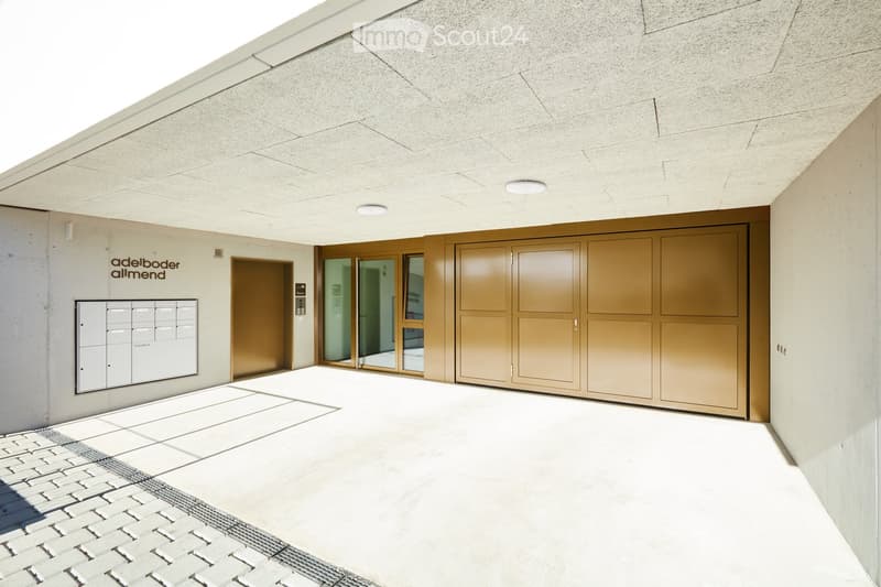 Modern building entrance with a raised ground floor, featuring a large garage door, mailboxes, and a sleek design with concrete and glass elements.