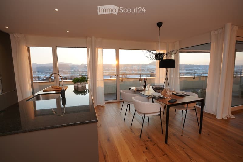 Modern kitchen with wooden flooring, large windows offering a scenic view, a sink with a modern faucet, a dining table with chairs, and decorative items including a bowl, wine bottle, and potted plants.
