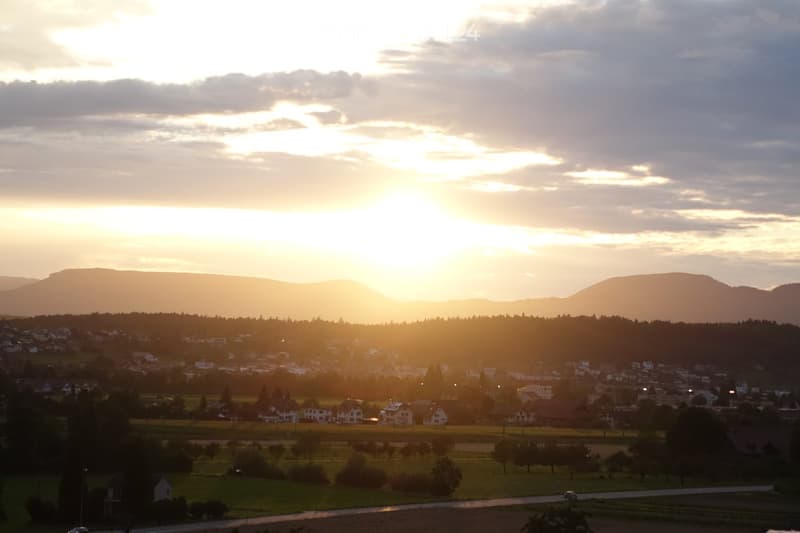 A panoramic view of a town at sunset, with houses, trees, and distant mountains silhouetted against a golden sky.