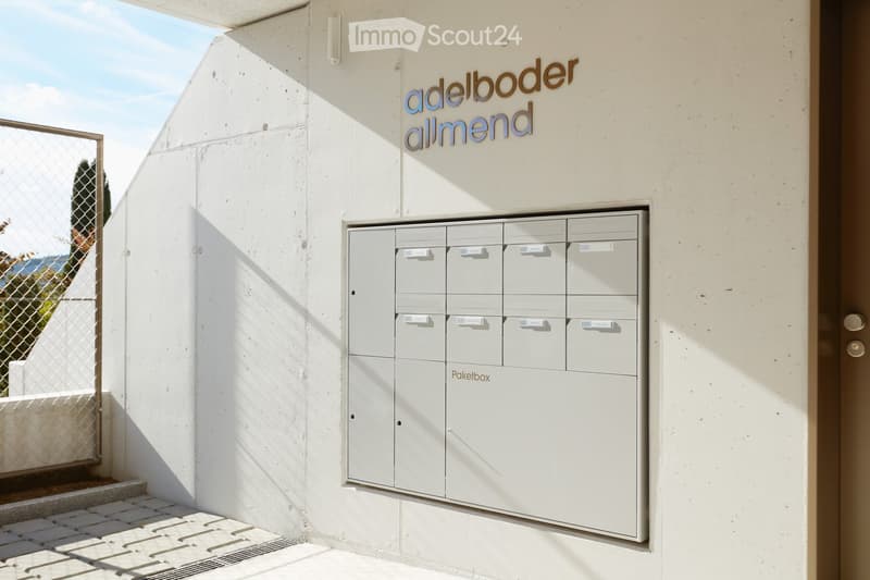Modern building entrance with mailboxes labeled 'Paketbox' and a sign reading 'adelboder allmend'.