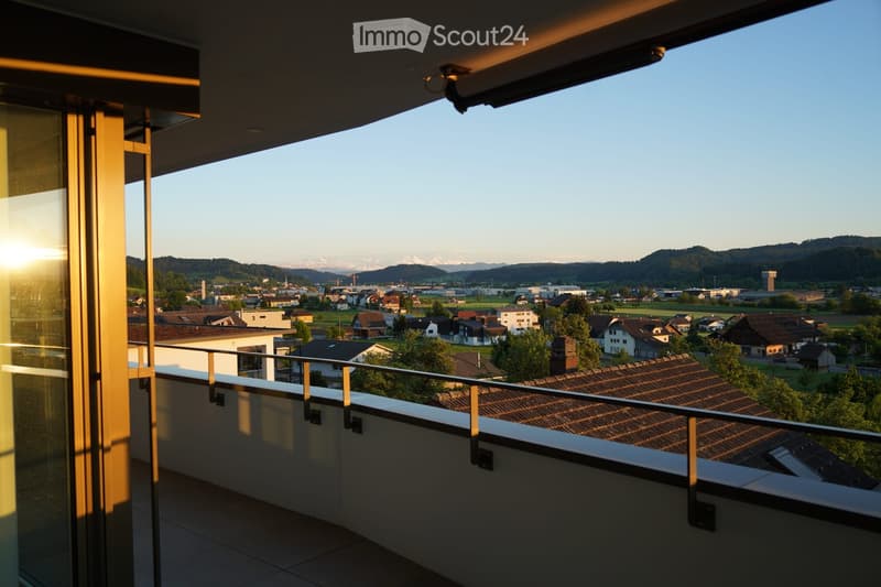 Balcony with a metal railing, overlooking a town with houses, fields, and distant mountains during sunset.