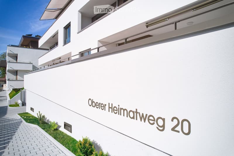 Modern white building with clean lines, multiple levels, balconies, and a sign reading "Oberer Heimatweg 20".