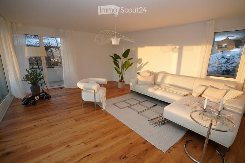 Modern living room with white sofa, glass coffee table, white armchair, floor lamp, and potted plants. Wooden flooring and large windows with sheer curtains.