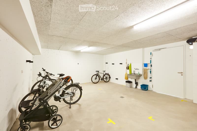 Empty room with white walls and concrete floor, featuring bicycles, a stroller, a sink, and cleaning supplies.