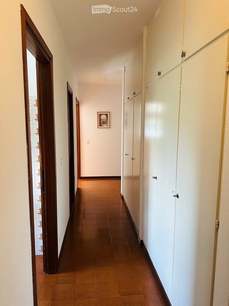 A narrow hallway with tiled flooring, white walls, and several closed doors. A small framed picture hangs on one wall.