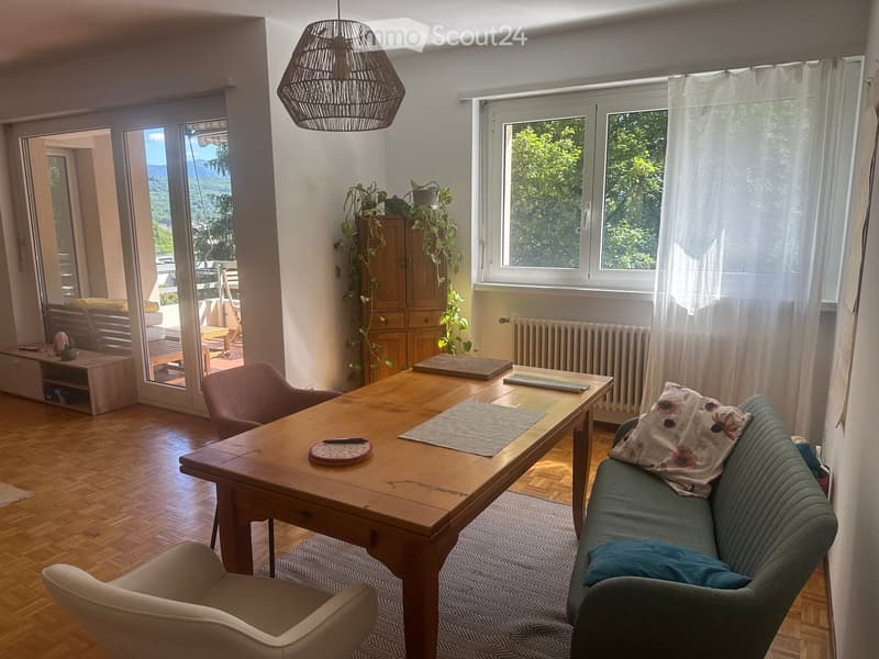 Bright living room with wooden flooring, large windows, and a balcony access. Features a sofa, wooden table, chairs, and a potted plant.