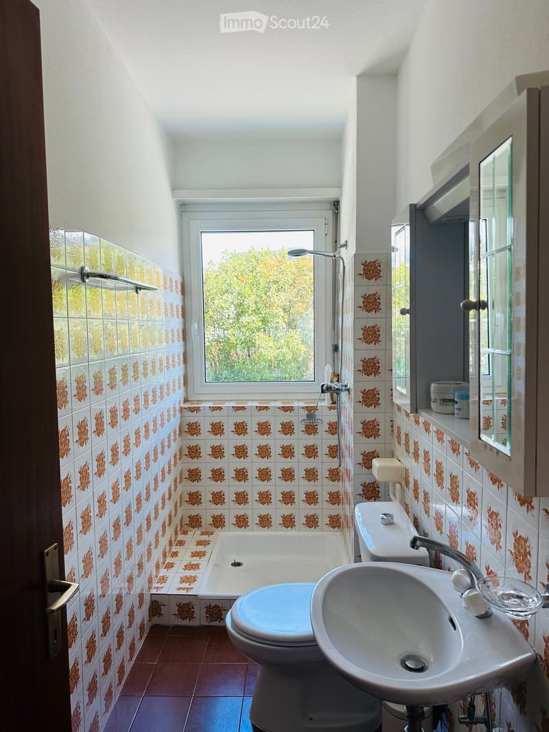Bathroom with floral-patterned tiles, a bathtub, toilet, sink, mirror, and a window offering a view of greenery.